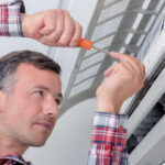HVAC technician in a plaid shirt servicing a wall-mounted mini-split air conditioning unit with a screwdriver.